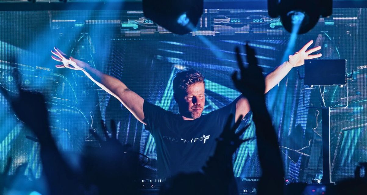 Ferry Corsten DJing to a packed crowd in a club with blue lighting. Everyone, including Ferry, has their hands in the air.