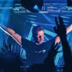 Ferry Corsten DJing to a packed crowd in a club with blue lighting. Everyone, including Ferry, has their hands in the air.