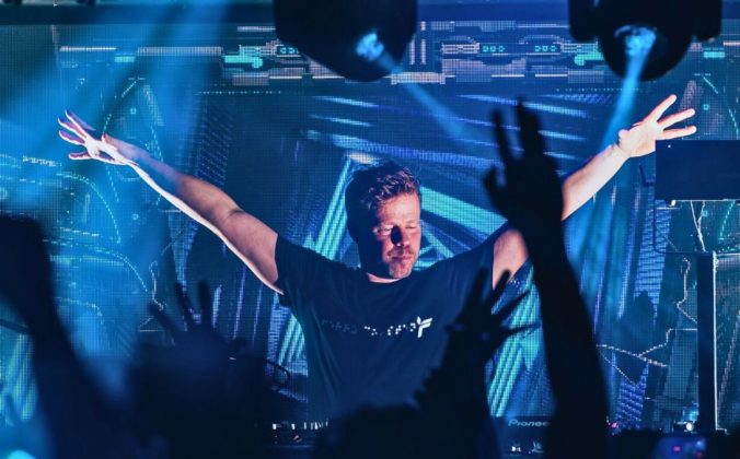 Ferry Corsten DJing to a packed crowd in a club with blue lighting. Everyone, including Ferry, has their hands in the air.