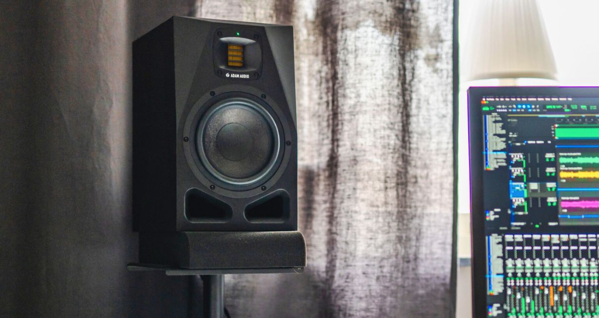 A black Adam Audio A7V studio monitor mounted on a stand, with a computer screen displaying music production software in the background.