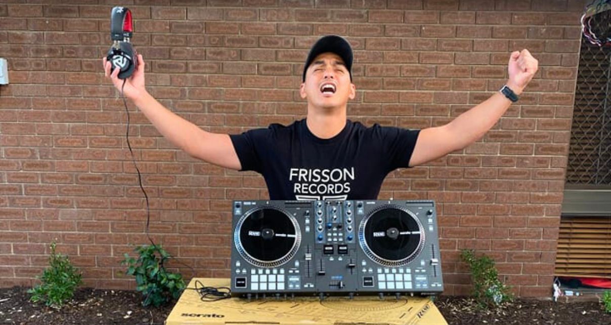 A DJ stands outdoors, in front of a brick wall and behind a propped-up Rane One DJ controller. He's closing his eyes, holding a pair of headphones, and raising his arms in celebration.