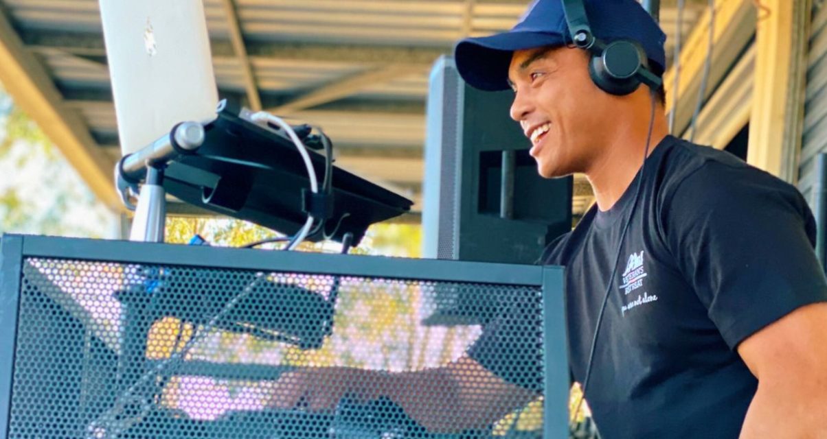 Side view of a DJ playing music behind a booth. He's wearing a black t-shirt and headphones and smiling at the crowd.
