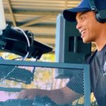 Student Spotlight: From Military Compound To Packed Dancefloors Side view of a DJ playing music behind a booth. He's wearing a black t-shirt and headphones and smiling at the crowd.