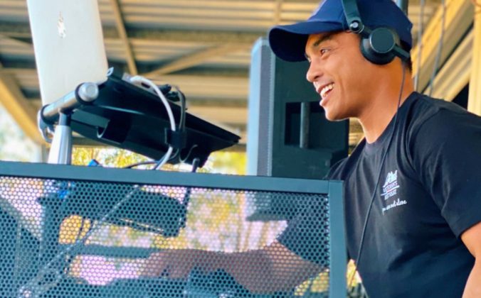 Side view of a DJ playing music behind a booth. He's wearing a black t-shirt and headphones and smiling at the crowd.