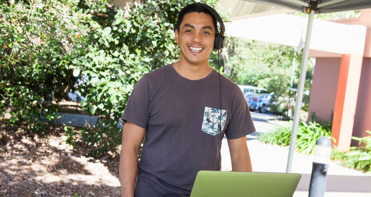 A DJ wearing a t-shirt and headphones smiles at the camera from behind an outdoor booth.
