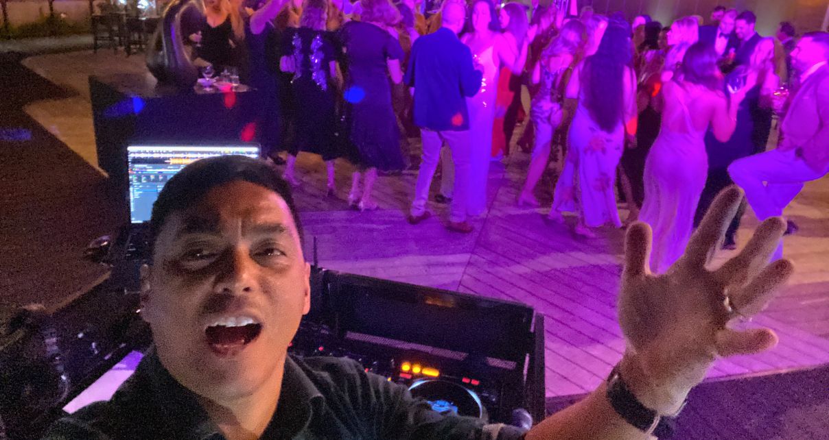 A DJ taking a selfie from behind the booth. A packed dancefloor is behind him under purple and pink lighting.
