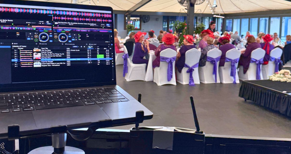 Photo taken from a DJ booth. On the left is a silver laptop displaying DJ software. In the background is a formal reception.