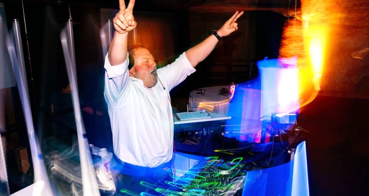 DJ Michael Gerth stands behind a lively booth, surrounded by lights. He's wearing a formal white button up shirt, rolled at the sleeves, headphones, and a watch. He's throwing both hands up in the air with peace signs, cheering to a crowd off screen.
