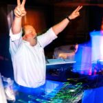 DJ Michael Gerth stands behind a lively booth, surrounded by lights. He's wearing a formal white button up shirt, rolled at the sleeves, headphones, and a watch. He's throwing both hands up in the air with peace signs, cheering to a crowd off screen.
