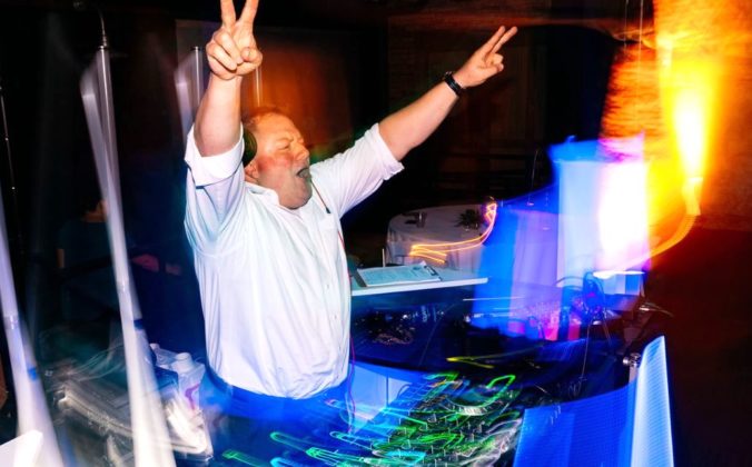 DJ Michael Gerth stands behind a lively booth, surrounded by lights. He's wearing a formal white button up shirt, rolled at the sleeves, headphones, and a watch. He's throwing both hands up in the air with peace signs, cheering to a crowd off screen.