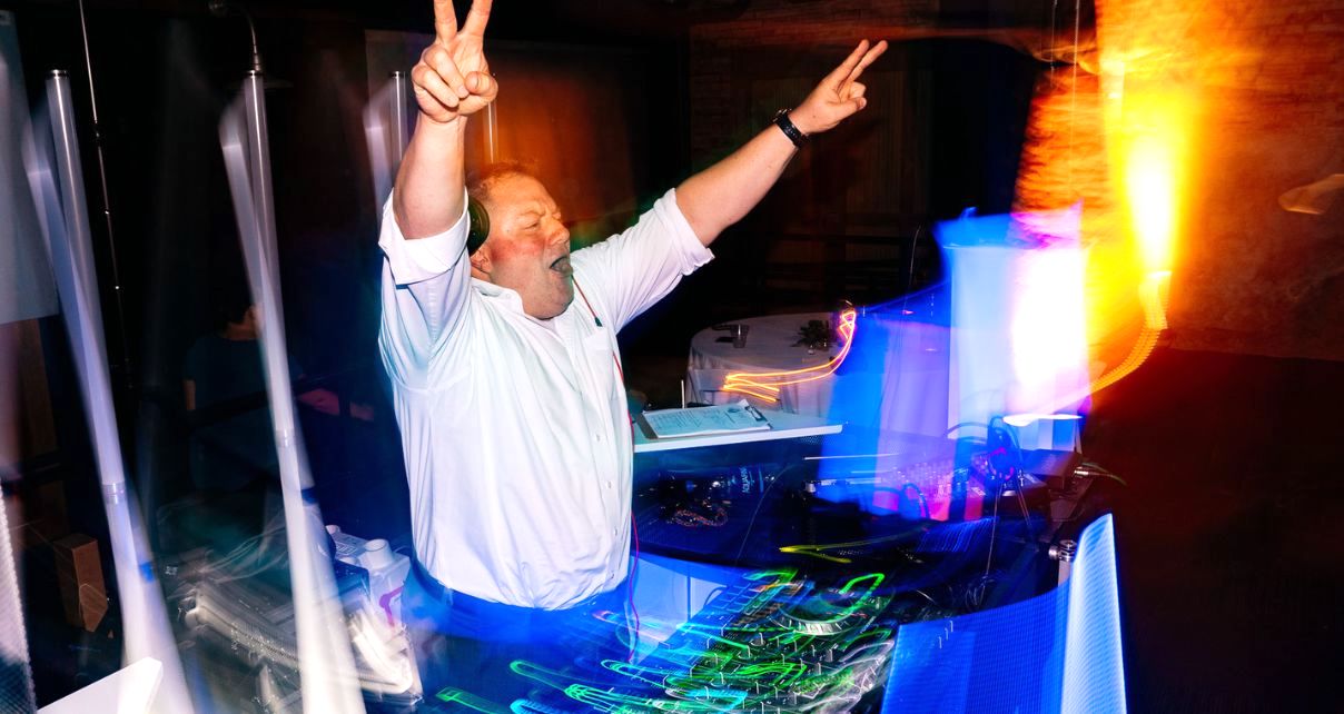 DJ Michael Gerth stands behind a lively booth, surrounded by lights. He's wearing a formal white button up shirt, rolled at the sleeves, headphones, and a watch. He's throwing both hands up in the air with peace signs, cheering to a crowd off screen.