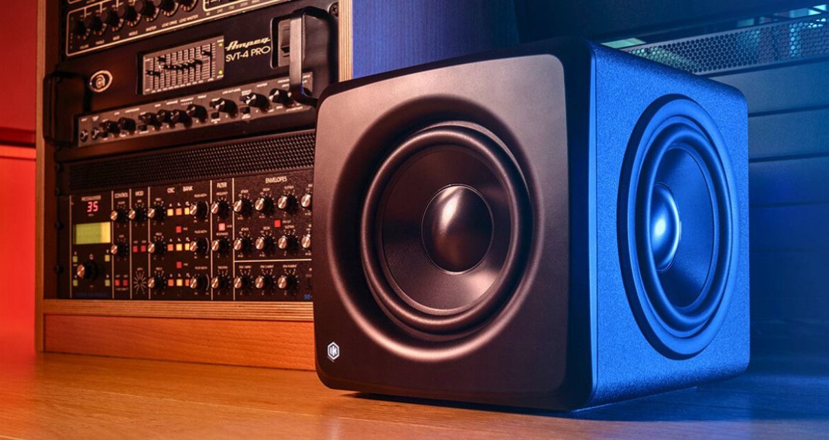 A single black iLoud Compact Studio Subwoofer sits on a wooden floor next to a rack of audio equipment.