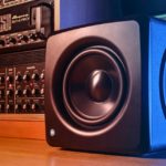 A single black iLoud Compact Studio Subwoofer sits on a wooden floor next to a rack of audio equipment.