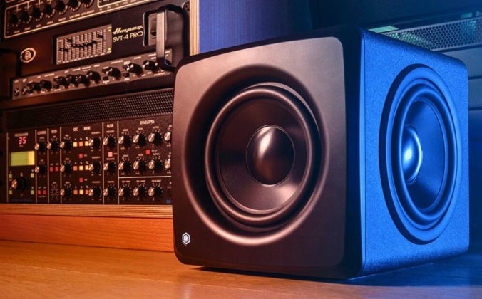 A single black iLoud Compact Studio Subwoofer sits on a wooden floor next to a rack of audio equipment.