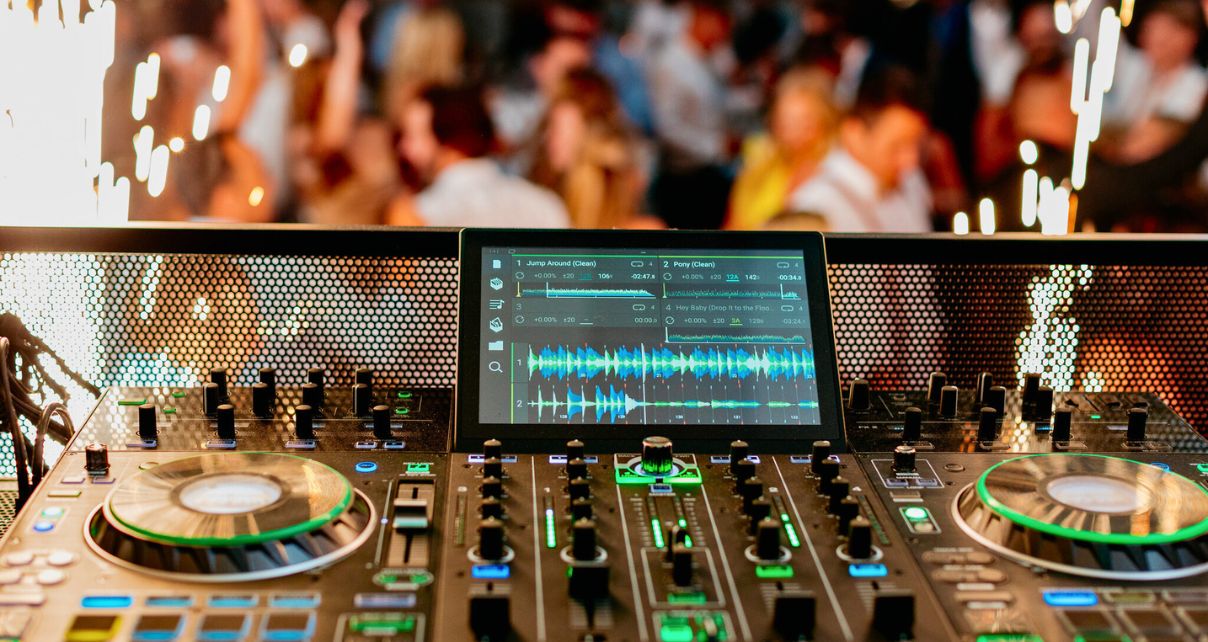 A Denon DJ system displaying vibrant waveforms and controls, set against a lively dance floor illuminated by sparkles.