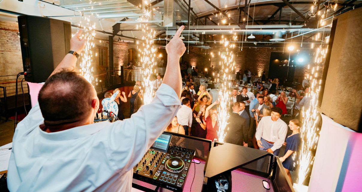 A DJ performs at a lively event, surrounded by dancing guests and sparkling pyrotechnics, creating a festive atmosphere.