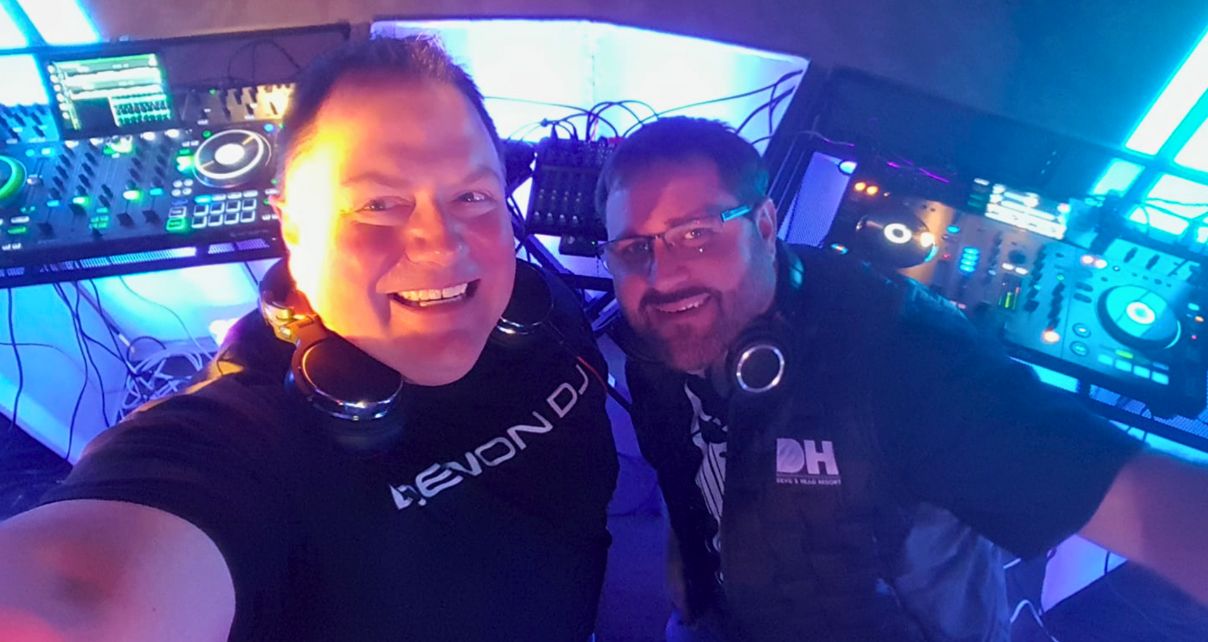 Two DJs posing for a selfie in front of a vibrant DJ setup with colourful lights and mixing equipment.