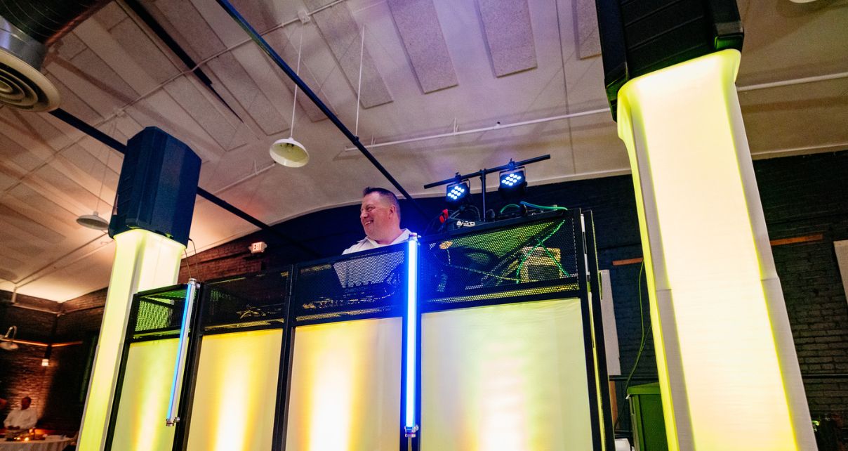 A DJ booth illuminated by yellow lights, with DJ equipment and large speakers, set in a stylish venue. The DJ smiles from behind the booth.