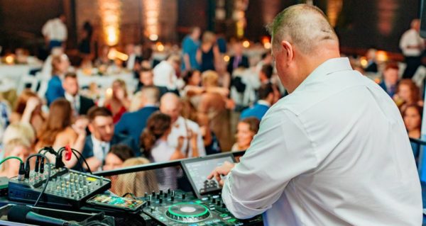 A DJ in a white dress shirt mixes music at a lively event, surrounded by guests enjoying the celebration and dancing in the background.