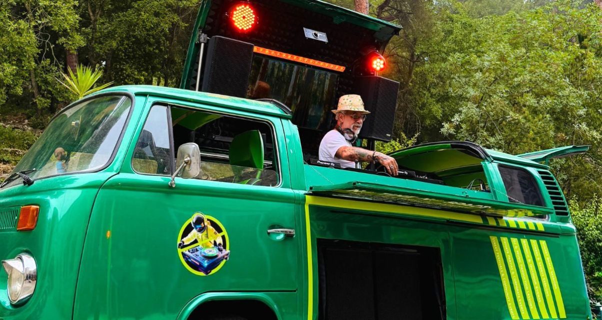 A person stands behind DJ gear in a custom-built green and yellow 1978 VW campervan. He's wearing a tan brimmed hat, sunglasses, a light t-shirt, and headphones while adjusting something on the DJ mixer. Behind him are two red lights and speakers.
