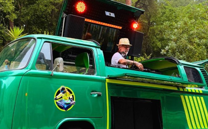 A person stands behind DJ gear in a custom-built green and yellow 1978 VW campervan. He's wearing a tan brimmed hat, sunglasses, a light t-shirt, and headphones while adjusting something on the DJ mixer. Behind him are two red lights and speakers.