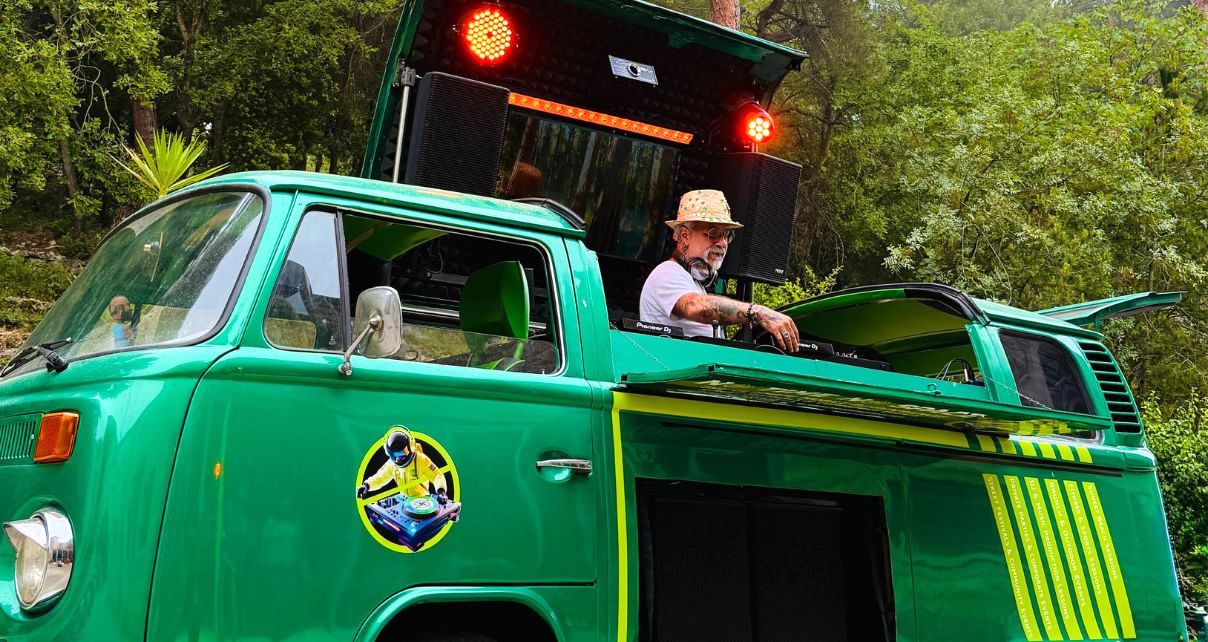 A person stands behind DJ gear in a custom-built green and yellow 1978 VW campervan. He's wearing a tan brimmed hat, sunglasses, a light t-shirt, and headphones while adjusting something on the DJ mixer. Behind him are two red lights and speakers.