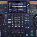 Top-down view of a black AlphaTheta XDJ-AZ standalone DJ system set on a wooden desk. The room is well-lit, and a person hands are resting on either side of the unit.