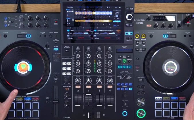 Top-down view of a black AlphaTheta XDJ-AZ standalone DJ system set on a wooden desk. The room is well-lit, and a person hands are resting on either side of the unit.