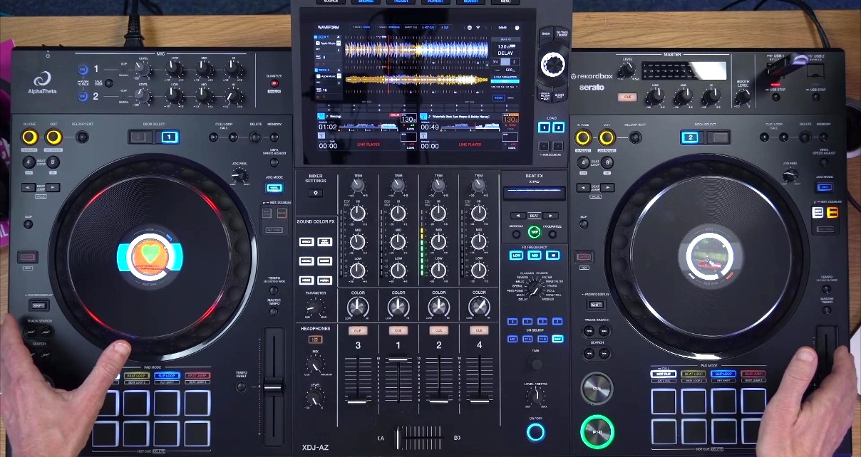 Top-down view of a black AlphaTheta XDJ-AZ standalone DJ system set on a wooden desk. The room is well-lit, and a person hands are resting on either side of the unit.