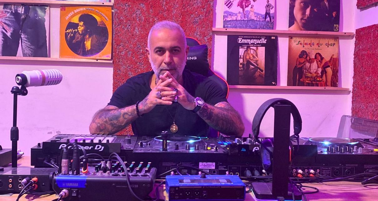 A photo of Jihad Zein, a man in his 50s with grey hair and a beard, wearing a black t-shirt, a necklace, rings, and a watch. His arms are covered in tattoos. His hands clasp together as he stares thoughtfully into the camera. Behind him are shelves on a wall holding vinyl records. In front of him is a lot of DJ gear sitting on a desk. 