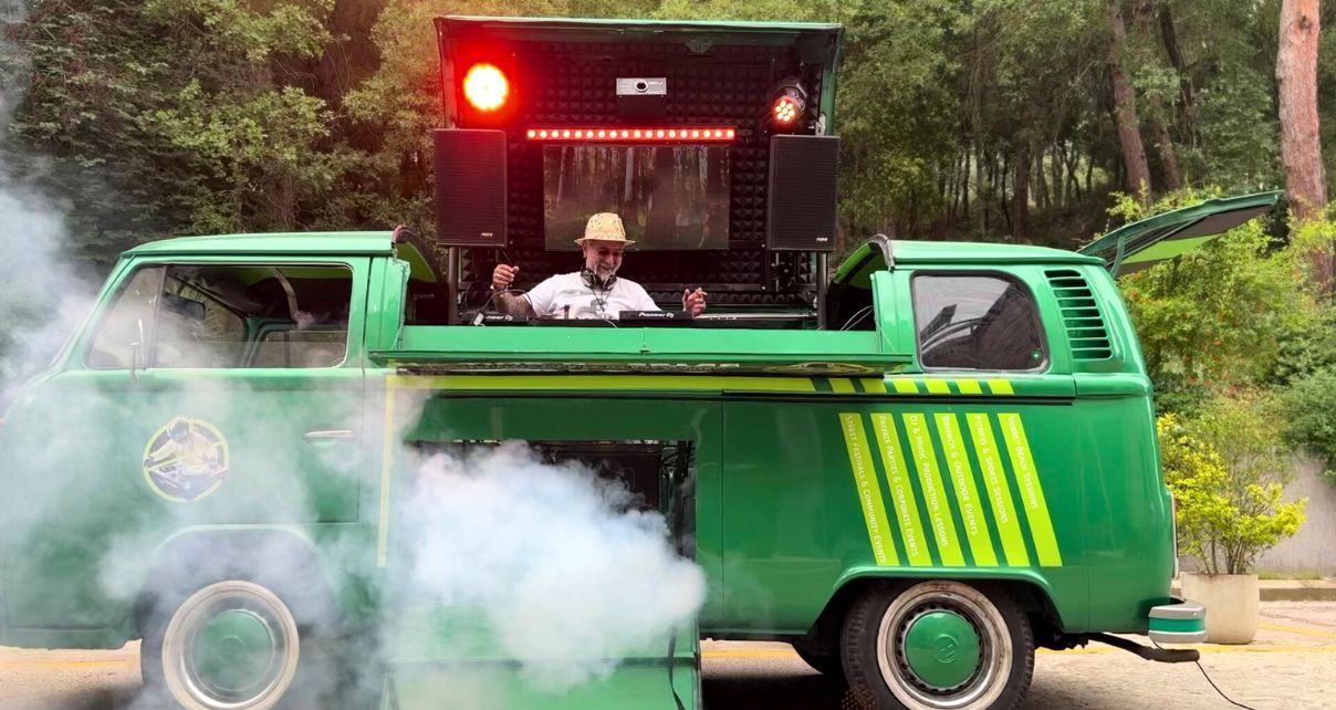 A person stands behind DJ gear in a custom-built green and yellow 1978 VW campervan. He's wearing a tan brimmed hat, sunglasses, a light t-shirt, and headphones while laughing and raising his arms. Behind him are two red lights and speakers. There's also a lot of smoke coming from the side of the van. 