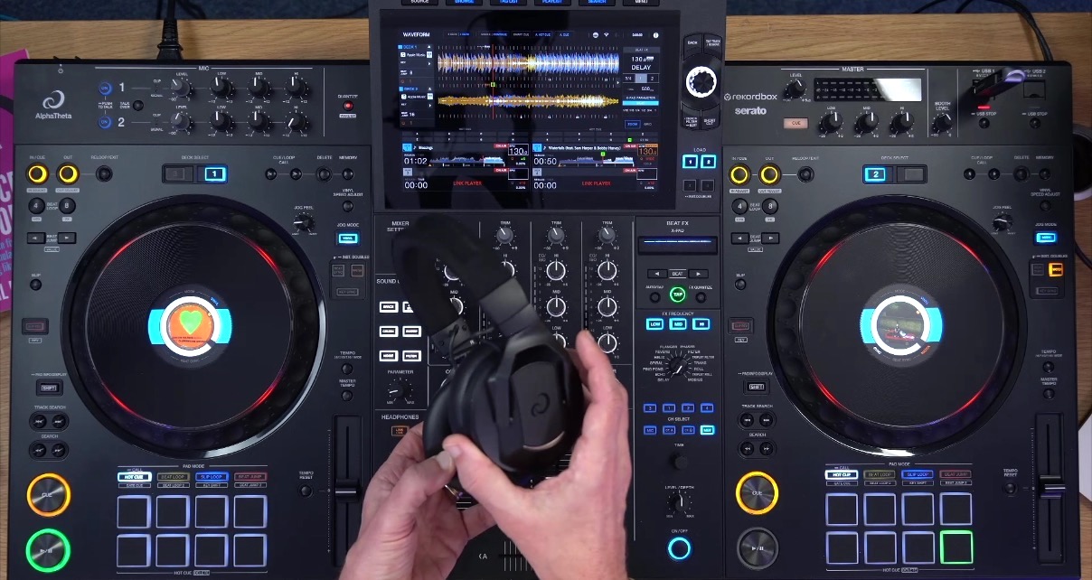 Top-down view of an XDJ-AZ standalone DJ system in a well-lit room. A person is holding a pair of HDJ-F10 DJ headphones with both hands over the unit.