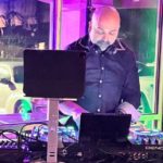 Student Spotlight: From South Bronx "Franken-rig" to Multi-Genre Mastery A DJ wearing a denim button up shirt and headphones around the neck looks down at a Denon DJ standalone system. There's a window behind him showing cars in a parking lot. The lighting inside the room is green and purple.