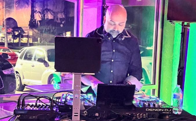 A DJ wearing a denim button up shirt and headphones around the neck looks down at a Denon DJ standalone system. There's a window behind him showing cars in a parking lot. The lighting inside the room is green and purple.