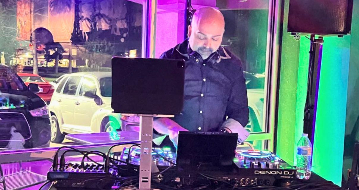 A DJ wearing a denim button up shirt and headphones around the neck looks down at a Denon DJ standalone system. There's a window behind him showing cars in a parking lot. The lighting inside the room is green and purple.