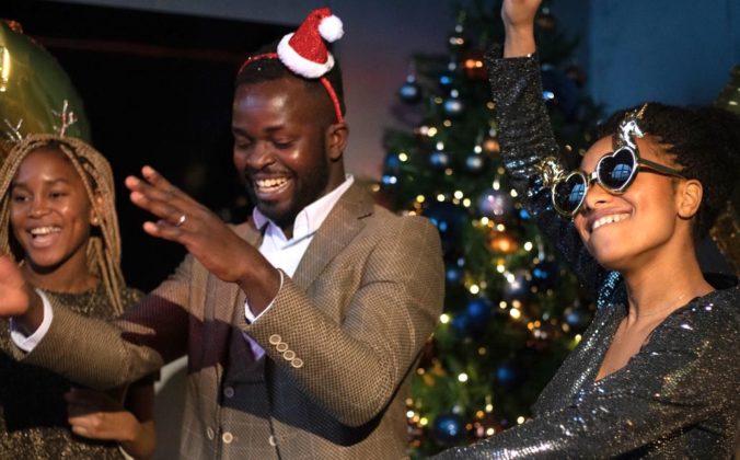 Three people dance and laugh in front of a Christmas tree. One is wearing a tiny santa hat and another is wearing heart sunglasses with unicorns on it.