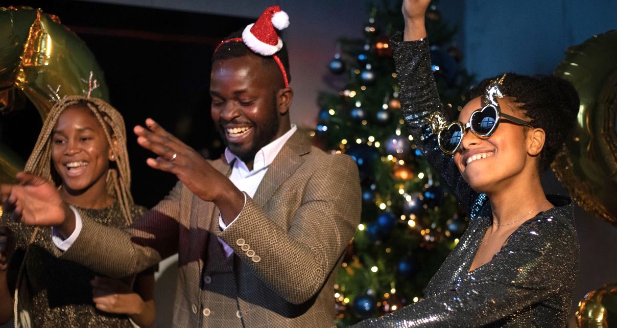 Three people dance and laugh in front of a Christmas tree. One is wearing a tiny santa hat and another is wearing heart sunglasses with unicorns on it.