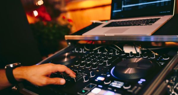 A slightly angled close-up photo of someone mixing on a DJ controller. A laptop is perched above and in the background. The person's hand is adjusting the volume faders. 