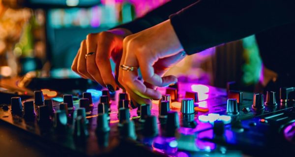 Close-up on a DJ's hands mixing songs on a controller. They appear to be wearing formal clothes. The colourful purple, blue, and green lighting reflects off of the controller. 
