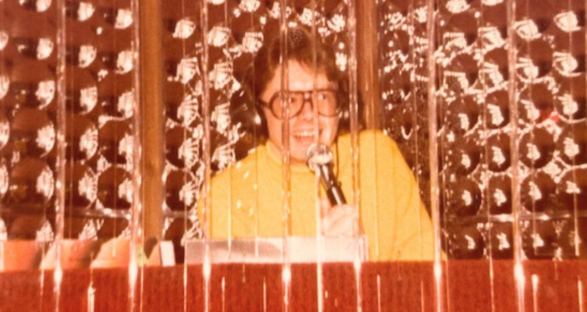 A photo of David Franks. He has longer, light brown hair, big glasses, and is wearing headphones and a light orange turtleneck. He's standing behind a DJ booth and holding up a microphone to speak.