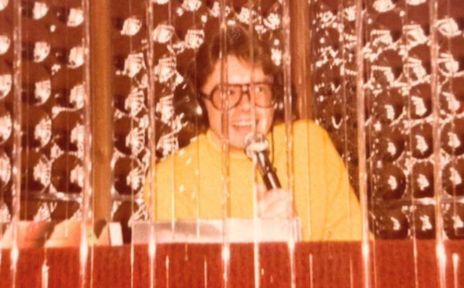 A photo of David Franks. He has longer, light brown hair, big glasses, and is wearing headphones and a light orange turtleneck. He's standing behind a DJ booth and holding up a microphone to speak.