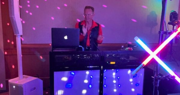 Photo of David Franks, a white man in his 70s. He's wearing a red button-up shirt underneath a black vest and has short hair and glasses. He appears to be clapping along to music. He's standing behind a laptop and DJ gear, surrounded by party lighting.