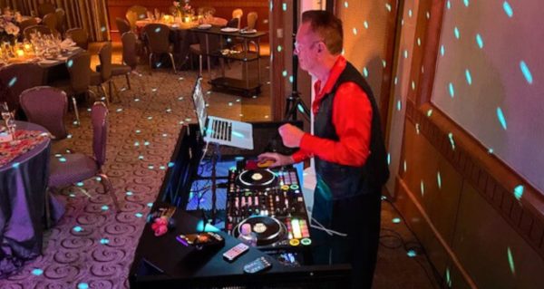 A side profile photo of David Franks, a white man in his 70s. He's wearing a red button-up shirt underneath a black vest and has short hair and glasses. He's standing behind a DJ booth that includes a controller and laptop. There are various empty dinner tables in front of the booth.