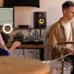 12 Non-DJ Skills That Will Help You Become a Better DJ Two musicians collaborate in a studio. On the left, a person with headphones plays an electric guitar at the computer, while the person on the right plays the drums.