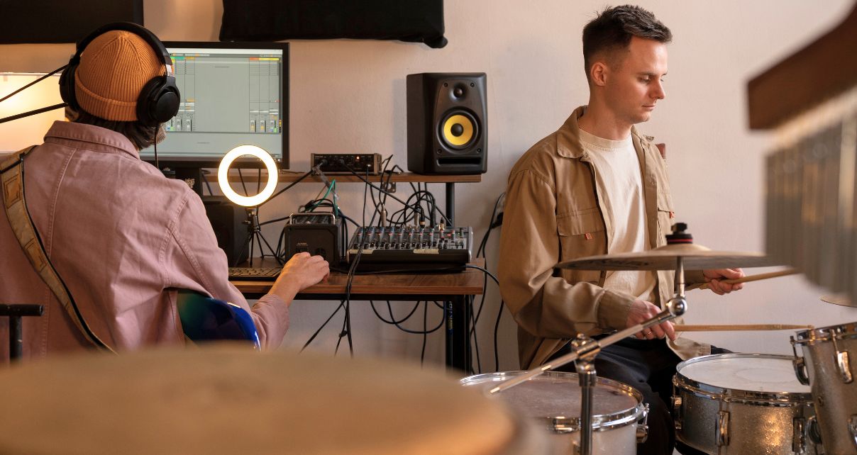 Two musicians collaborate in a studio. On the left, a person with headphones plays an electric guitar at the computer, while the person on the right plays the drums.