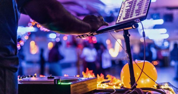 Photo taken from behind a DJ booth. A DJ searches for a track on their laptop. Turntables and digital vinyl sit on the table in front of them. A blurry dancefloor is in the background. 