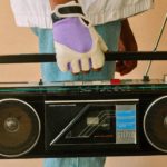 A person stands, facing sideways, wearing purple and white gloves. They're holding an old black boombox.