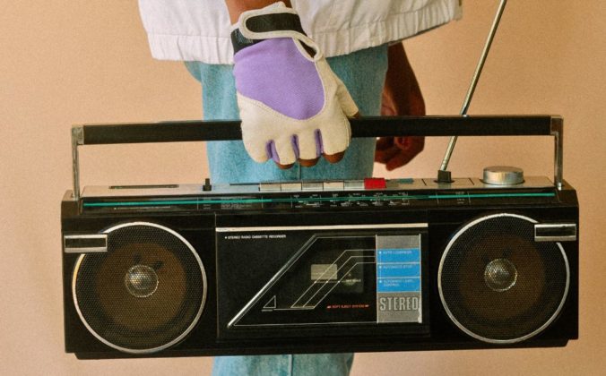 A person stands, facing sideways, wearing purple and white gloves. They're holding an old black boombox.