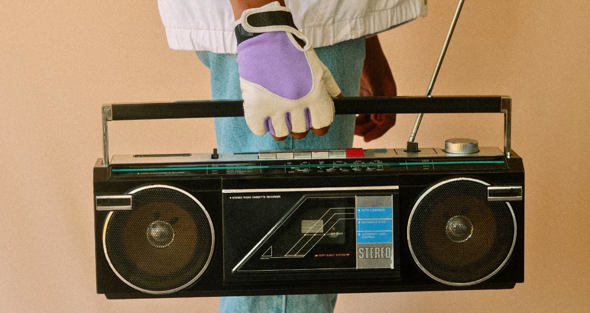 A person stands, facing sideways, wearing purple and white gloves. They're holding an old black boombox.