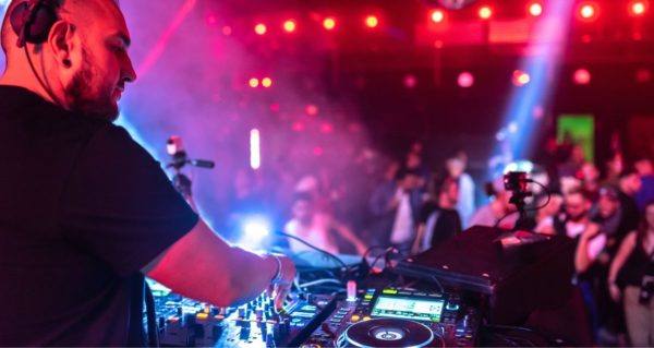 A club DJ adjusts the mixer from behind a pro booth. He looks onward as a blurry crowd dances in the background. 